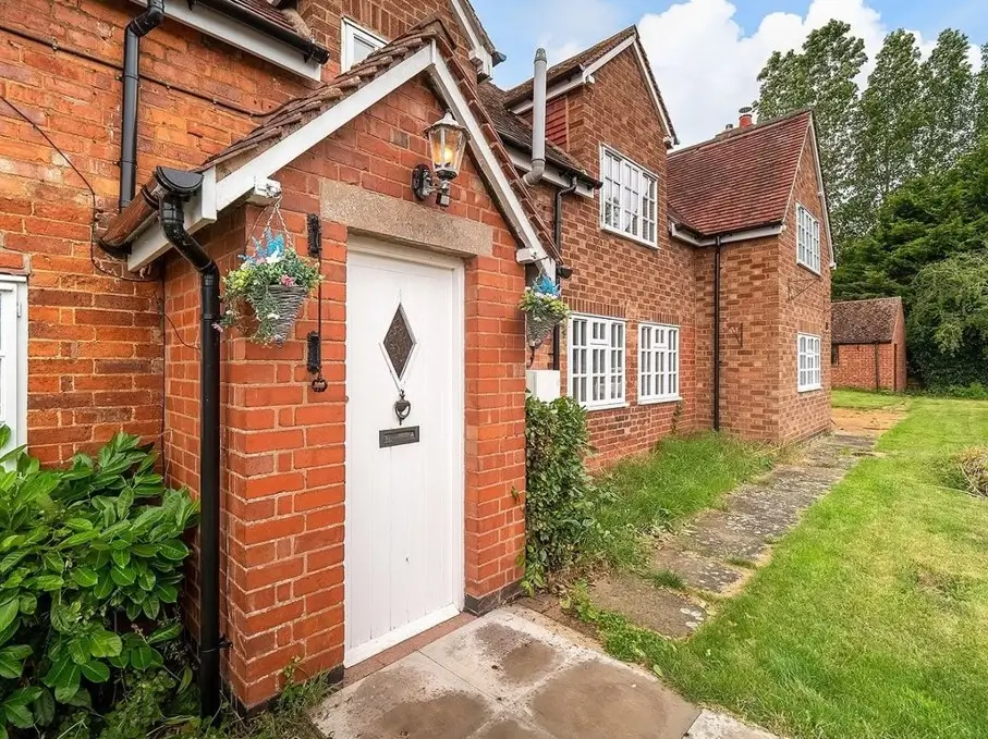 White Trim Cottage — White framing wraps this brick cottage — a leaded plank door, gabled porch, and white cottage windows against warm red brick and a stone surround.