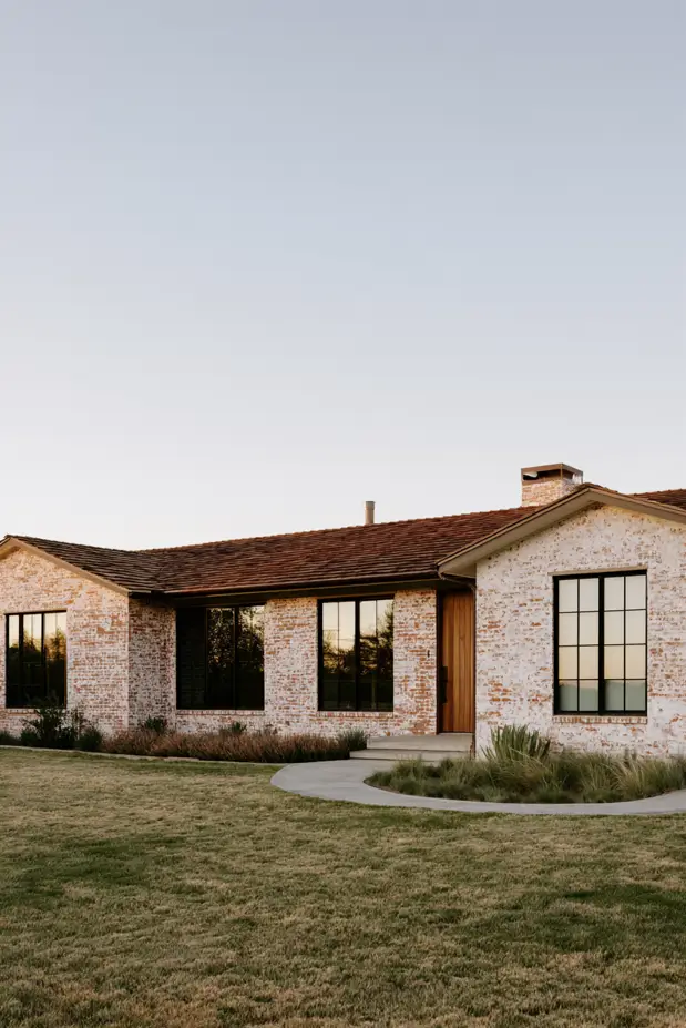 Low single-story ranch in limewashed brick with a clay-tile roof, black-framed windows, wood door, and grasses at the base