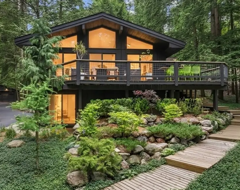 Black chalet-style A-frame house exterior with glowing windows above a boulder and fern rock garden