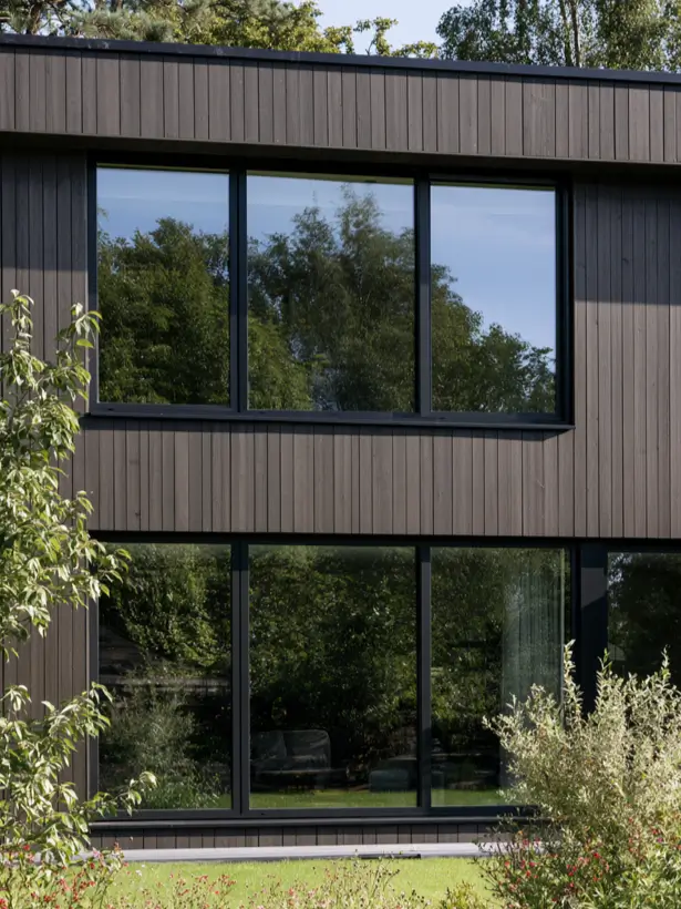 Dark stained vertical timber house exterior with large black-framed windows reflecting the garden