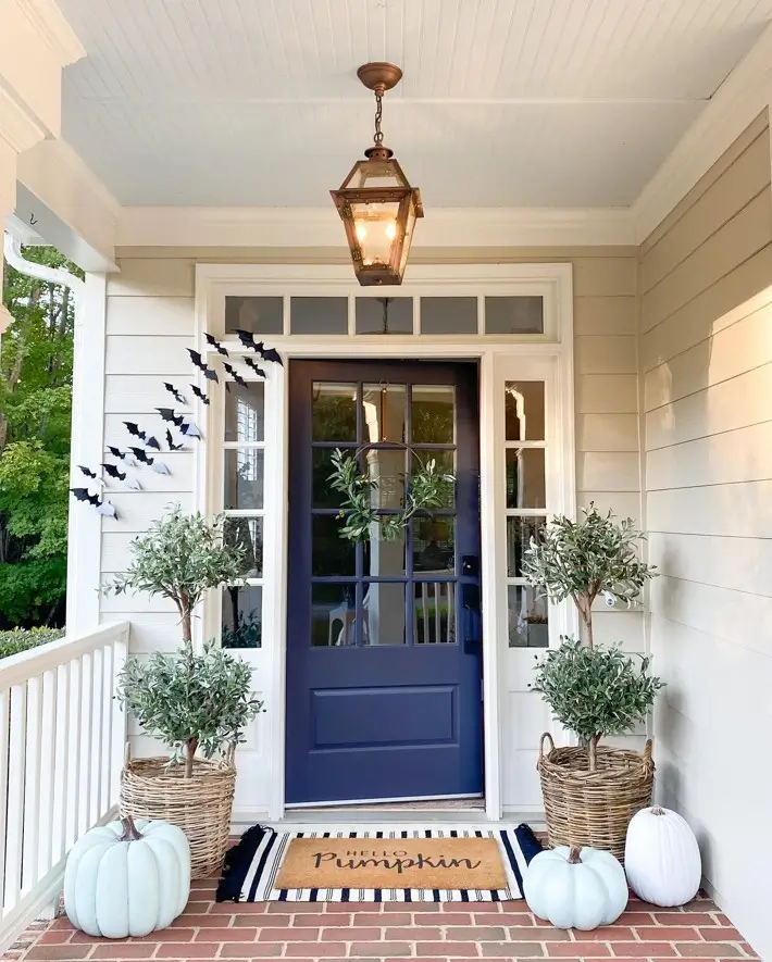 Navy blue glass-paned front door with white trim, flanked by olive topiaries in wicker baskets on a brick porch.