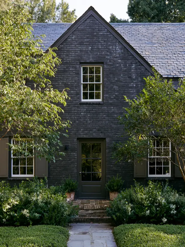 Charcoal painted brick cottage exterior with olive shutters, white sash windows and boxwood hedging