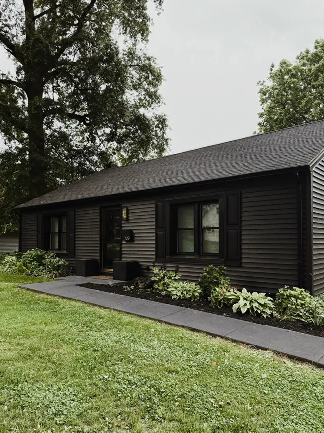 Black ranch house exterior with dark lap siding, matching shutters, brass house numbers and hosta plantings