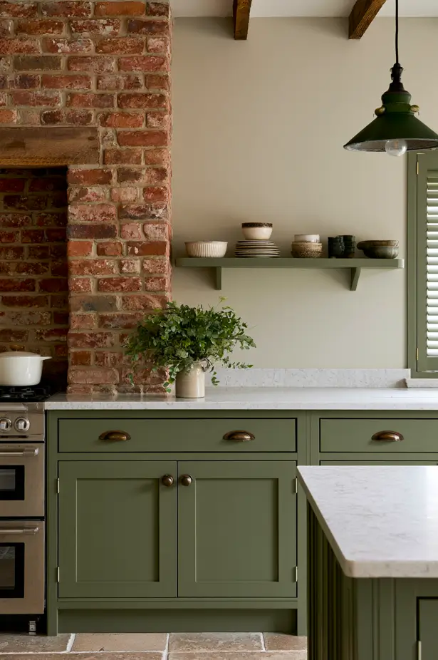 Olive Green Kitchen — Olive-green Shaker cabinets and a matching shelf meet an exposed brick chimney breast, softened by brass handles and a wood mantel beam.