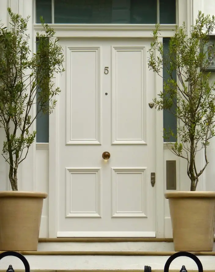 Creamy white paneled front door with brass centre knob and house number, flanked by symmetrical olive trees in terracotta pots.