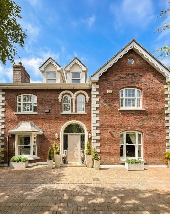 Classic Cream Trim — Off-white sash windows, stone quoins, and carved bargeboards trim this traditional red brick house, with a soft greige door under an arched stone entry.
